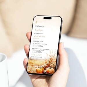 May include: A smartphone displays a baptism invitation with a fall theme. The invitation features gold text and a cross icon at the top. The background shows a field with pumpkins and flowers. The text includes the name Annabele Rose and event details.