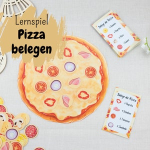 Op de afbeelding: Een pizzabelagspel voor kinderen. Het spel bevat een groot pizzabord, kleine pizzabelagstukjes en kaarten met instructies voor het maken van een pizza. De kaarten zijn in het Duits en zeggen "Belege die Pizza", wat "Bedek de pizza" betekent.