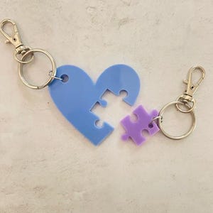 Op de afbeelding: Een blauwe hartvormige sleutelhanger met een ontbrekend puzzelstukje, en een paarse puzzelstukje sleutelhanger. Beide sleutelhangers zijn bevestigd aan zilveren sleutelringen.