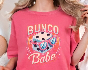 May include: A watermelon-colored t-shirt with a graphic that says "Bunco Babe." The graphic features a die wearing sunglasses. The shirt is a short-sleeved, crew neck style.