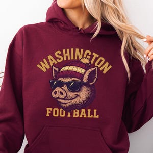 May include: Maroon hoodie with a front graphic of a pig wearing sunglasses and a beanie. The text "WASHINGTON FOOTBALL" is arched above the pig. The hoodie has a kangaroo pocket and a drawstring hood.