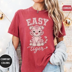 May include: A crimson short-sleeve t-shirt featuring a graphic of a seated tiger cub with a patterned texture. The text "EASY" appears in block letters above the tiger, and the word "tiger" is written in cursive script below with a small heart.
