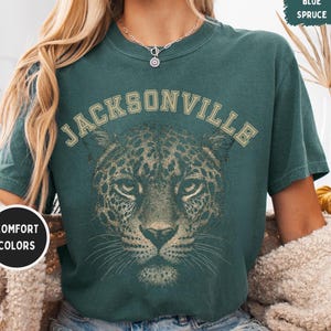 Puede incluir: Camiseta de color verde azulado con la palabra "JACKSONVILLE" arqueada sobre la cara de un leopardo. La cara del leopardo está detallada con manchas y bigotes. La camiseta también tiene las palabras "COMFORT COLORS" en un círculo blanco y "BLUE SPRUCE" en un rectángulo azul.