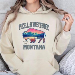Könnte beinhalten: Ein beiger Hoodie mit einer Grafik eines Bison-Silhouetts mit einem Sonnenuntergang und Bergen darin. Der Text "Yellowstone National Park Montana" ist unter der Grafik gedruckt.