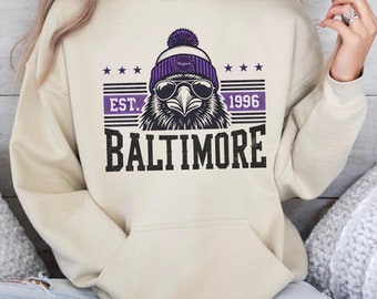 May include: A cream coloured hoodie with a purple and black graphic of a bird wearing a purple and white beanie and sunglasses. The graphic also includes the text "EST. 1996 BALTIMORE".