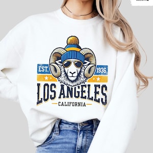 Puede incluir: Sudadera blanca con un gráfico de estilo vintage de un carnero con un gorro azul y amarillo y gafas de sol. El gráfico dice "EST. 1936 LOS ANGELES CALIFORNIA".