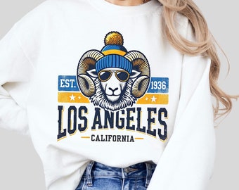 May include: White sweatshirt featuring a vintage-style graphic of a ram wearing a blue and yellow beanie and sunglasses. The graphic reads "EST. 1936 LOS ANGELES CALIFORNIA".