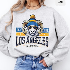 Puede incluir: Sudadera gris con un gráfico de un carnero con un gorro azul y amarillo y gafas de sol. El gráfico también incluye el texto "EST. 1936 LOS ANGELES CALIFORNIA".