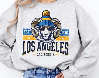 May include: Grey sweatshirt featuring a graphic of a ram wearing a blue and yellow beanie and sunglasses. The graphic also includes the text "EST. 1936 LOS ANGELES CALIFORNIA".