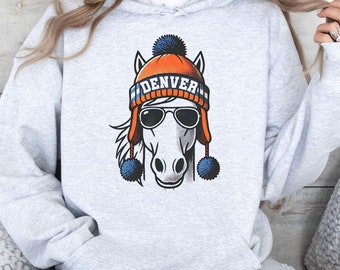 Puede incluir: Una sudadera gris con un caballo blanco que lleva un gorro azul y naranja con la palabra "DENVER" en él. El caballo lleva gafas de sol y tiene pompones azules en las orejas.