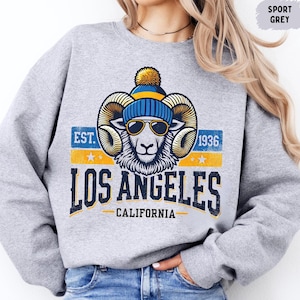 Puede incluir: Una sudadera gris con un gráfico de un carnero que lleva un gorro azul y amarillo y gafas de sol. El gráfico también incluye el texto "EST. 1936 LOS ANGELES CALIFORNIA".
