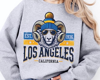 May include: A grey sweatshirt featuring a graphic of a ram wearing a blue and yellow beanie and sunglasses. The graphic also includes the text "EST. 1936 LOS ANGELES CALIFORNIA".