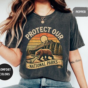 May include: A dark gray t-shirt with a vintage-style graphic of a bear in a forest with the text "Protect Our National Parks".