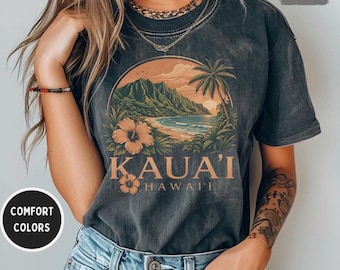 Puede incluir: Camiseta gris oscuro con una escena de playa tropical y el texto "KAUAI HAWAII". El diseño incluye una playa, montañas, palmeras y flores de hibisco. La camiseta está etiquetada como "Comfort Colors" y "Pepper".