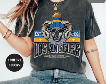 Könnte beinhalten: Ein graues T-Shirt mit einer Vintage-Grafik eines Widders, der eine blaue und gelbe Mütze und eine Sonnenbrille trägt. Die Grafik enthält auch den Text "EST. 1936 LOS ANGELES CALIFORNIA".