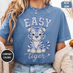 May include: A blue jean coloured short-sleeve t-shirt featuring a graphic of a tiger with a blue and white plaid and floral patchwork pattern. The text EASY is printed in a block font above the tiger, and the word tiger is in script below.
