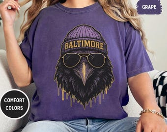 Puede incluir: Una camiseta morada con un gráfico de un cuervo con gorro y gafas de sol. La palabra "BALTIMORE" está en el gorro. La camiseta es de color confort y el color es uva.