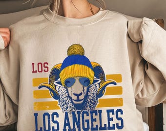 May include: A beige sweatshirt with a graphic print of a cartoon ram wearing sunglasses and a blue and yellow beanie. The text "LOS ANGELES" is printed below the ram.