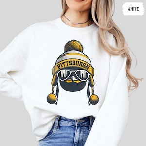 Könnte beinhalten: Weißer Pullover mit einem Aufdruck eines Pinguins, der einen schwarzen und gelben Hut mit dem Wort "PITTSBURGH" trägt. Der Pinguin trägt eine Sonnenbrille.