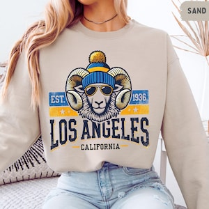 Puede incluir: Una sudadera beige con un gráfico de un carnero que lleva un gorro azul y amarillo y gafas de sol. El gráfico también incluye el texto "EST. 1936 LOS ANGELES CALIFORNIA".