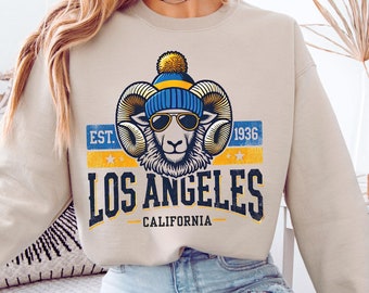 May include: A beige sweatshirt with a graphic of a ram wearing a blue and yellow beanie and sunglasses. The graphic also includes the text "EST. 1936 LOS ANGELES CALIFORNIA".