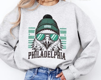 May include: A grey sweatshirt featuring a graphic of a cartoon eagle wearing a green beanie and sunglasses. The eagle is looking to the right and the text "PHILADELPHIA" is printed below the eagle. The sweatshirt is being modelled by a person wearing blue jeans.