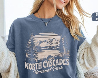 Może przedstawiać: Koszulka z dżinsu w kolorze niebieskim z grafiką przedstawiającą pasmo górskie i jezioro z napisem "North Cascades National Park" w kolorze kremowym.