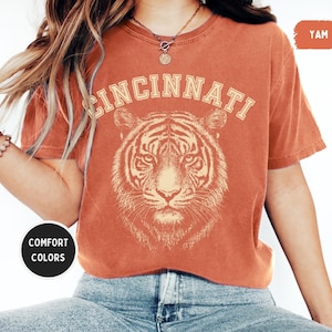 May include: Burnt orange t-shirt featuring the word "CINCINNATI" above a tiger graphic. The shirt is a comfort color and is paired with blue jeans. The shirt is a short-sleeved crew neck.