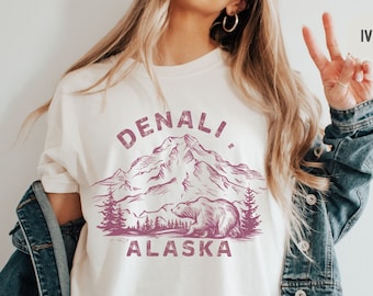 May include: A white t-shirt with a pink graphic of a mountain range and a bear with the text "Denali, Alaska". The model is wearing a denim jacket and has blonde hair.