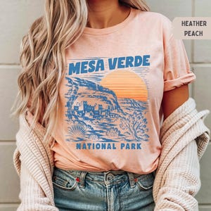May include: A heather peach t-shirt featuring a blue graphic print of Mesa Verde National Park. The design includes a landscape of cliff dwellings, desert vegetation, and a large orange sun. The text reads MESA VERDE NATIONAL PARK.