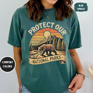 May include: A teal blue t-shirt with a vintage-style graphic of a bear in a forest scene. The graphic says "Protect Our National Parks".