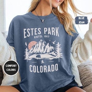 May include: A blue jean t-shirt with the text "Estes Park Estd. 1917 Colorado" and a graphic of a mountain range with a bear and a campfire. The shirt is available in comfort colors.