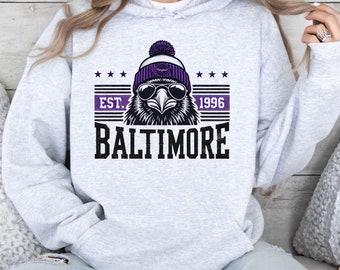 May include: A grey hoodie featuring a black and white graphic of a bird wearing a purple beanie and sunglasses. The graphic also includes the text "EST. 1996 BALTIMORE".
