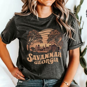 Puede incluir: Una camiseta de color gris oscuro jaspeado con un gr&aacute;fico de estilo vintage del horizonte de Savannah, Georgia. El gr&aacute;fico presenta una puesta de sol, &aacute;rboles y edificios. El texto "Savannah Georgia" est&aacute; impreso en una fuente retro.