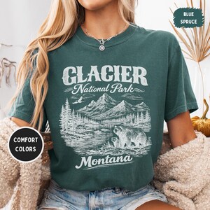以下が含まれることがあります： 氷河国立公園のグラフィックが描かれた、ティール色のTシャツ。山、木、クマが描かれています。「Glacier National Park Montana」の文字がヴィンテージスタイルで書かれています。シャツには「Comfort Colors」と「Blue Spruce」と書かれています。