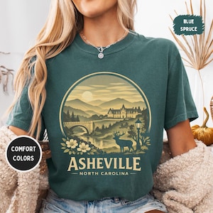 May include: A teal-colored t-shirt featuring a circular graphic of Asheville, North Carolina. The design includes a bridge, mountains, a deer, and the text "Asheville - North Carolina". The shirt also has the text "Comfort Colors" and "Blue Spruce".