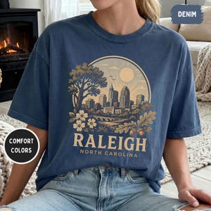 May include: A denim blue t-shirt featuring a circular graphic of the Raleigh, North Carolina skyline, with the text "RALEIGH NORTH CAROLINA". The design includes a tree, flowers, and a bridge. The shirt has short sleeves.