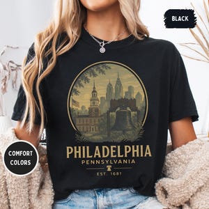 Könnte beinhalten: Schwarzes T-Shirt mit einem kreisförmigen Grafikdesign von Wahrzeichen von Philadelphia, darunter Independence Hall und die Liberty Bell. Der Text "PHILADELPHIA PENNSYLVANIA EST. 1681" ist darunter gedruckt. Das Shirt hat "COMFORT COLORS" und "BLACK" Etiketten.