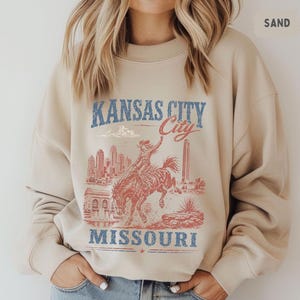 May include: Sand-colored sweatshirt featuring a distressed graphic of Kansas City, Missouri. The design includes a cowboy riding a bucking bronco, city skyline, and the text "Kansas City" and "Missouri" in blue and red.