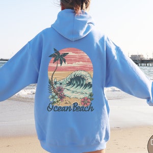 Op de afbeelding: Lichtblauwe hoodie met een retro graphic op de rug. Het ontwerp toont een palmboom, een oceaangolf en de woorden "Ocean Beach". De graphic bevat roze, blauwe en groene bloemaccenten. De hoodie is lichtblauw.