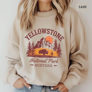 May include: A beige sweatshirt with a vintage-style graphic of Yellowstone National Park, Montana. The graphic features a mountain range, a setting sun, pine trees, and a bison.