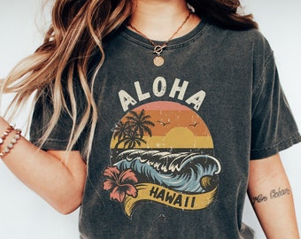 May include: A dark gray cropped t-shirt with a vintage-style graphic of a sunset over a beach with palm trees and waves. The graphic says "Aloha Hawaii" in a retro font.