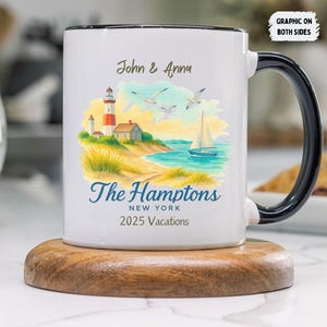 May include: White ceramic mug with a black handle and rim, featuring a coastal scene with a lighthouse, sailboat, and the text "The Hamptons, New York, 2025 Vacations." The mug is on a wooden coaster.