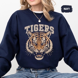Puede incluir: Una sudadera azul marino de cuello redondo con un gráfico de una cara de tigre con los dientes al descubierto y la palabra TIGERS impresa en una fuente universitaria de color beige sobre el tigre. Tiene mangas largas y puños acanalados.