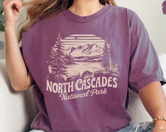 Może przedstawiać: Fioletowy t-shirt z grafiką w stylu vintage przedstawiającą górską scenerię i tekst "NORTH CASCADES National Park".