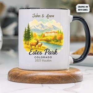 May include: White ceramic mug with a black handle and rim, featuring a colorful illustration of a mountain landscape, deer, and the text "Estes Park Colorado 2025 Vacation". The mug also has the names "John & Anna".