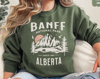 May include: A green sweatshirt with a white graphic design featuring a mountain range, a bear, and the text "Banff National Park - Since 1885 - Alberta".