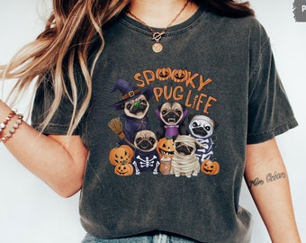 Puede incluir: Camiseta gris oscuro con un diseño de Halloween. El diseño presenta varios pugs disfrazados, incluyendo una bruja, un vampiro y una momia, con el texto "Spooky Pug Life" en naranja.