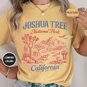 May include: Mustard-colored t-shirt with a graphic of Joshua Tree National Park. The design features a desert landscape with Joshua trees, mountains, a coyote, and the text "Joshua Tree National Park California". The shirt has a relaxed fit.