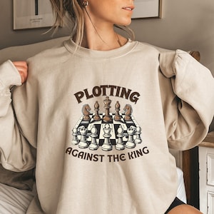 May include: A beige sweatshirt with a brown graphic of a chessboard and the text "Plotting Against The King".